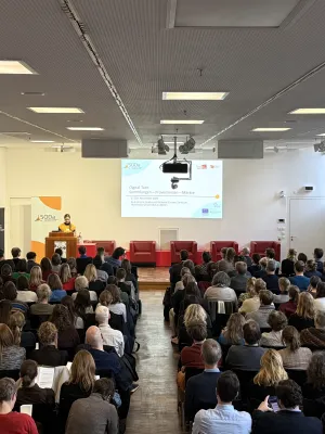 Im Auditorium des Grimm-Zentrum sitzen 150 Konferenzteilnehmer auf Stühlen. Sie hören dem Einführungsvortrag von Gabriele Zöllner zu, die auf einer Bühne hinter einem Podest steht und die Konferenz eröffnet. 